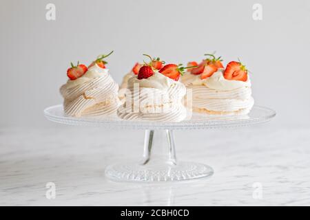 Mini Pavlova meringue nests with whipped cream and homegrown organic fresh strawberries. The mini meringue desserts are presented  on a glass cake sta Stock Photo