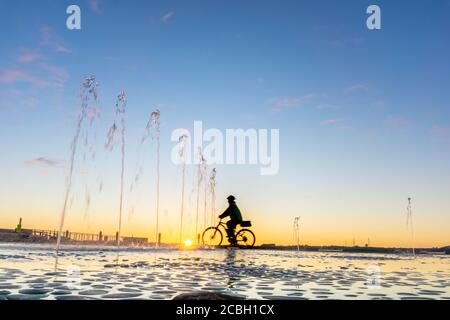 Water feature with spraying water on Tauranga Strand waterfront at sunrise with clear blue sky and blurred silhouette of cyclist passing in distance,