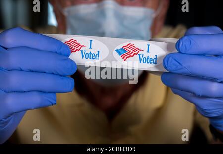 Election volunteer with face mask and medical gloves holding roll of I Voted buttons with USA Flag ready for in person voters in Presidential election Stock Photo