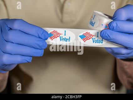 Hands in medical gloves holding roll of I Voted buttons with USA Flag ready for in person voters in Presidential election Stock Photo