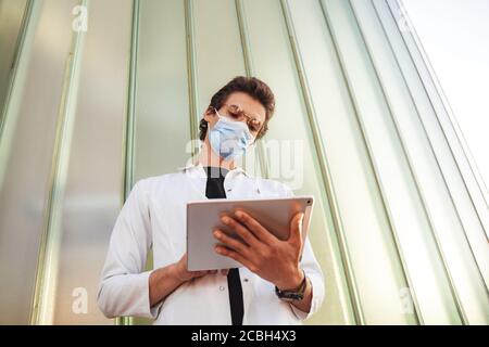 Caucasian young male doctor wearing face mask while using digital tablet against wall during COVID-19 outbreak Stock Photo