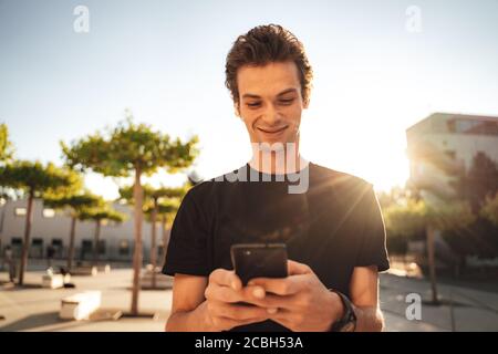 Handsome Caucasian university student smiling while using smartphone in city on sunny day Stock Photo