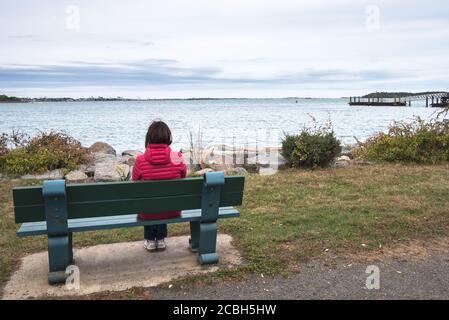Lonely woman sitting on a bench in front of a bay on a Cloudy Autumn Day. Concept of loneliness. Stock Photo