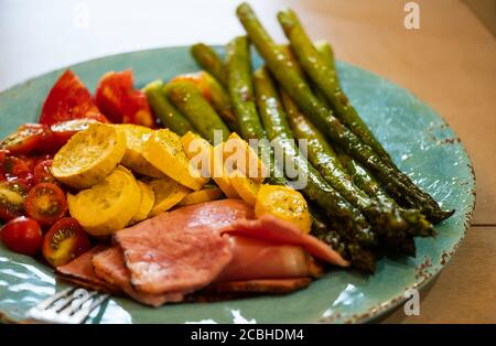 Seasoned asparagus, yellow squash, and tomatoes drizzled with olive oil and broiled. Served with ham slices on a turquoise dinner plate. Stock Photo