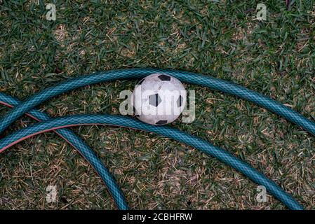 Top view of a dirty black and white football next to a green hose in a garden forming an interesting shape. Stock Photo