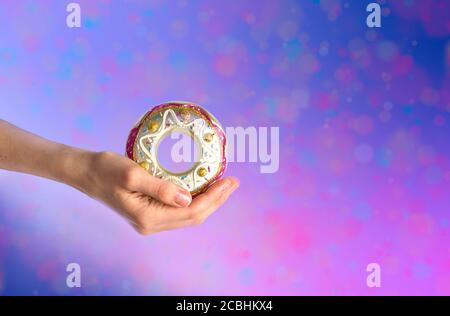 Woman hand gently holding Christmas toy - decorative colorful glass doughnut on multicolored pink-purple. Xmas invitation. Creative festive greeting c Stock Photo