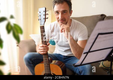 Smiling man with his acoustic guitar in hand looking straight ahead sitting on the sofa with music stand with songs at home Stock Photo