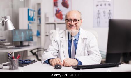 Mature doctor wearing eyeglasses and smiling in hospital cabinet sitting at desk. Stock Photo