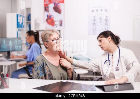 Doctor in hospital cabinet examining neck glands of senior patient. Mature woman with thyroid discomfort. Nurse in blue uniform working on computer. Stock Photo