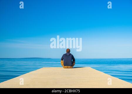 Man sitting on the edge of dock and meditating. Sea horizon at sunset, island od Pag, Adriatic sea, Croatia. Stock Photo