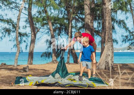 Camping people outdoor lifestyle tourists put up set up green grey campsite summer forest near lazur sea. Boy son helps father study mechanism of Stock Photo