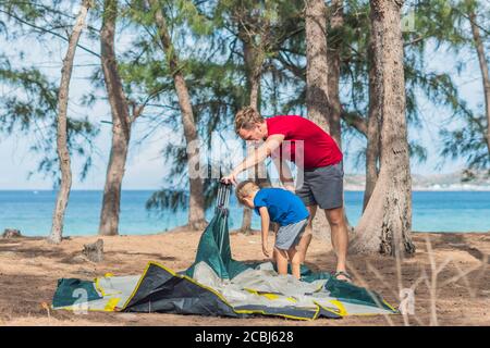 Camping people outdoor lifestyle tourists put up set up green grey campsite summer forest near lazur sea. Boy son helps father study mechanism of Stock Photo