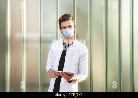 Caucasian young male doctor wearing face mask while using digital tablet against green wall during COVID-19 outbreak Stock Photo