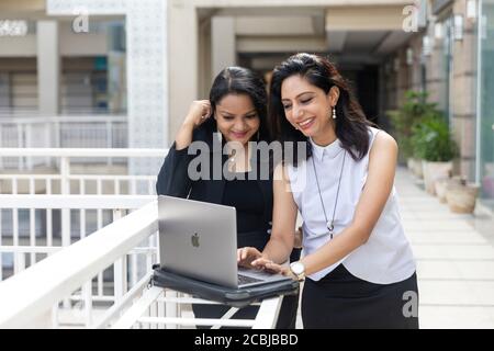 Two smiling  Indian women looking into a laptop in an urban corporate setting, Stock Photo