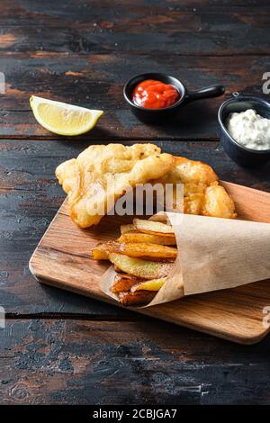 British Traditional Fish and chips with mashed peas, tartar sauce inn crumpled paper cone on wood chopping board dip and lemon - fried cod, french Stock Photo