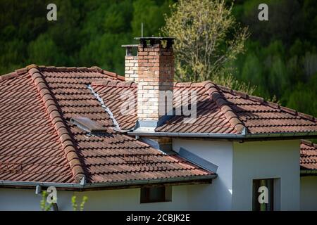 A fragment of a tiled roof with a chimney and windows in a new house in the woods in Bulgarian village. Stock Photo