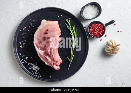 Raw Pork Loin chops in a black round plate on a white stone background with rosemary garlic peppercorns ingredients for grill top view Stock Photo