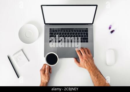 Programmer's workplace. A white table with a notebook, a computer mouse, a laptop, and men's hands holding a cup of espresso. Stock Photo