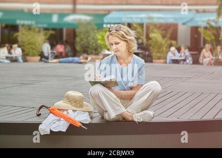Active caucasian senior woman about 60 years old is sitting in the public park and reading a book. White aged lady in casual and elegant cloth walks o Stock Photo