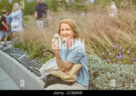 Charming white senior lady 62 years old is walking in the public park and sitting on the bench marked for social distance during coronavirus covid-19 Stock Photo
