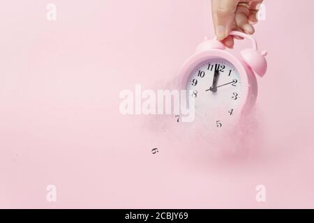 A hand holds alarm clock break down and dispersion to particle. A concept of the time. Light pink background. Concept Time is running up Stock Photo