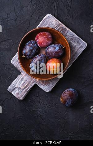 Fresh sweet plum fruits in brown wooden bowl on old cutting board, black textured background, top view Stock Photo