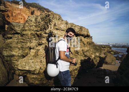 A young male photographer with a facemask holding a camera and a camera stand at the Chiclana beach, Spain Stock Photo