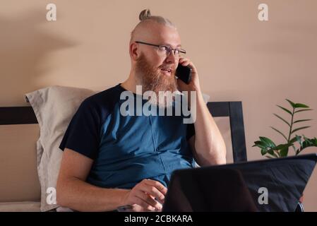 Middle aged businessman working from home talking on mobile phone while working on laptop. Smiling red bearded man solves work issues remotely Stock Photo