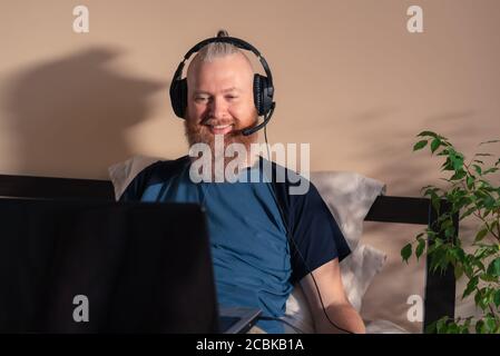 Smiling bearded man in headphones sitting in bed with laptop working remotely from home. Distance working and freelance work concept. Staying at home Stock Photo