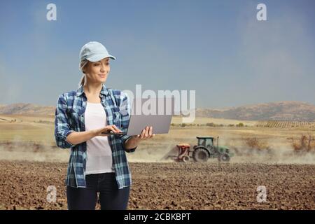 Young woman with a laptop computer standing on a field with a tractor operating on the soil Stock Photo