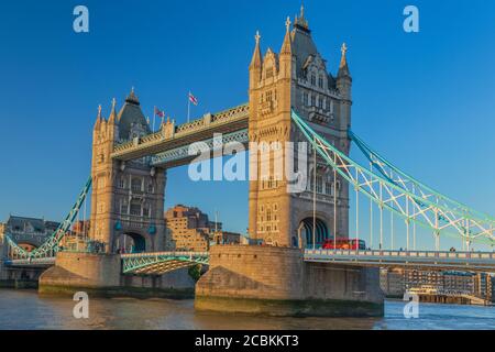 England, London, Tower Bridge in evening light viewed from the south bank of the River Thames with an iconic red London bus crossing the bridge. Stock Photo