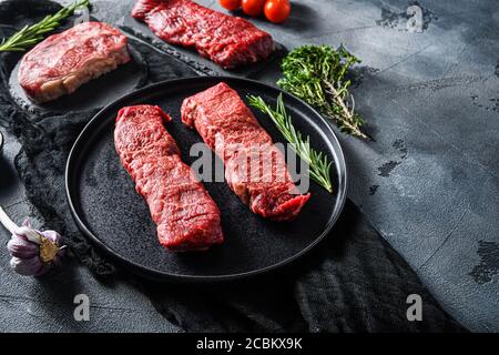 Raw denver, top blade, tri tip steak on a black plate and stone slate with seasonings, herbs grey concrete background. Side view close up selective Stock Photo