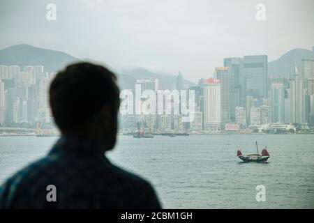 Over shoulder view of man looking out at chinese junk crossing Victoria harbour, Hong Kong, China Stock Photo