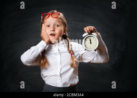 Back to school. Funny little girl schoolgirl with an alarm clock on the background of a black chalk board. The shocked child worries about the deadlin Stock Photo