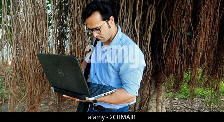 DISTRICT KATNI, INDIA - OCTOBER 23, 2019: An indian corporate man executive working on laptop around natural tree environment background. Stock Photo