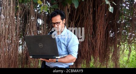 DISTRICT KATNI, INDIA - OCTOBER 23, 2019: An indian corporate smart boy working on laptop around natural tree root environment background. Stock Photo