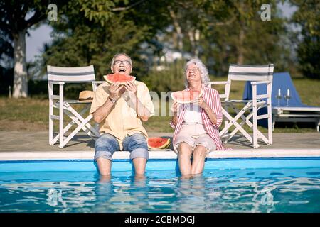 Elderly  couple  eating watermelon by the pool.Happy man and woman enjoy on summer holiday . Stock Photo