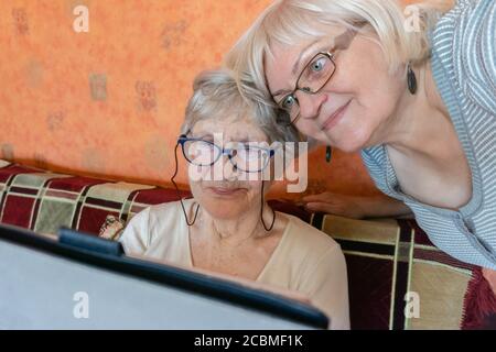 Old mother and daughter reading good news online inside. Woman teaching senior mother to use internet at home. Senior woman with her daughter looking Stock Photo