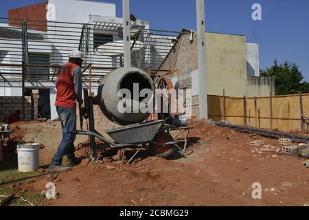 Man, worker in construction area, removing cement from mixer in wheelbarrow, for concrete under construction, in panoramic view Stock Photo