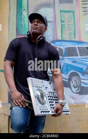 Cuban disc jockey with a modern dj turntable mixer equipment in front of a street painting wall art. Stock Photo