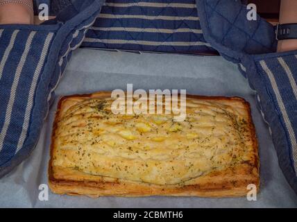 Woman’s hands in oven gloves holding an oven tray of greaseproof paper and a hot, flaky puff pastry, with salmon filling topped with cheese and dill. Stock Photo