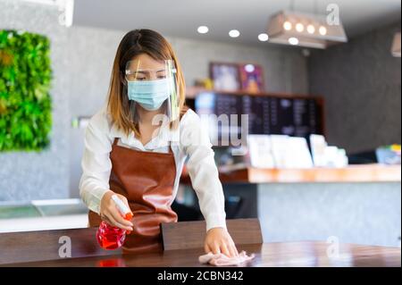 New normal startup small business Portrait of Asian woman barista wearing protection mask and face shield cleaning table in coffee shop while social d Stock Photo