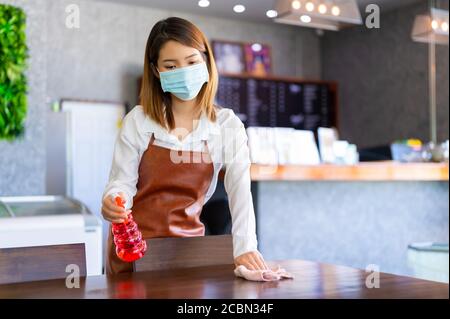 New normal startup small business Portrait of Asian woman barista wearing protection mask and face shield cleaning table in coffee shop while social d Stock Photo