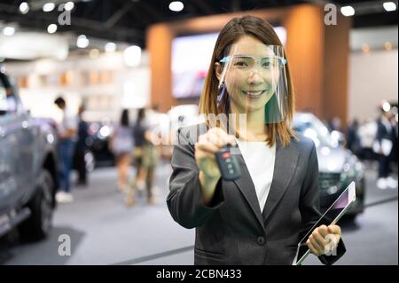 Asian car seller wearing face mask presents car and passing new car key to customer after purchased while coronavirus pandemic Stock Photo