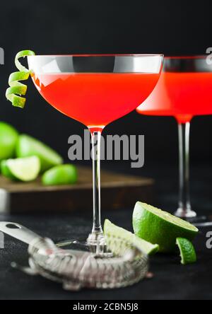 Cosmopolitan cocktail in modern crystal glasses with lime peel and fresh limes with strainer on black table background. Stock Photo