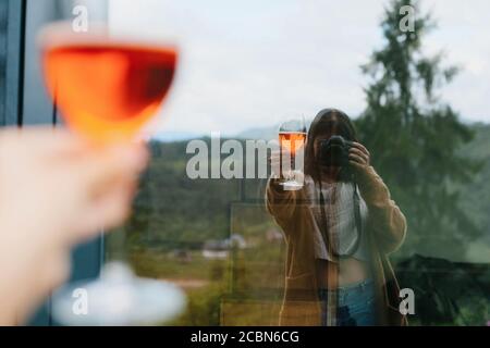 Woman holding aperol drink in hand and taking selfie in big window on background of mountains, summer vacation and resort. Woman cheering with delicio Stock Photo