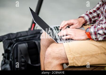 Close up of man hand typing on laptop keyboard while sitting on wooden bench in downtown. The male freelancer uses laptop and an electric scooter Stock Photo