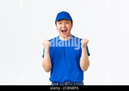 Excited and happy asian courier in blue uniform shouting yes and rejoicing, fist pump as celebrating great news, triumphing over white background Stock Photo
