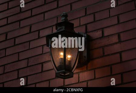 a black iron metal antique illuminated wall sconce on an exterior brick wall at night giving off a warm glowing light Stock Photo