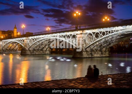 Young couple drinking sparkling wine by the Guadalquivir river and the Triana bridge, Seville, Spain Stock Photo